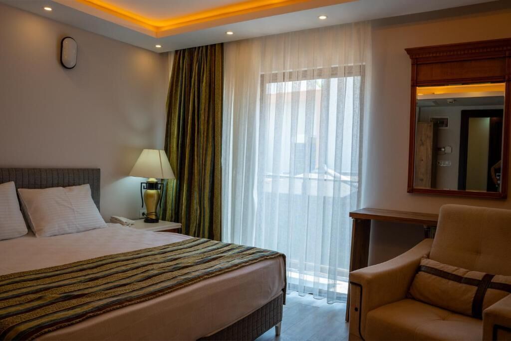 Habitación de hotel con una cama, una silla, una mesita de noche con lámpara, un espejo y una ventana con cortinas translúcidas.