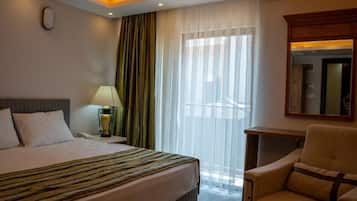 Habitación de hotel con una cama, una silla, una mesita de noche con lámpara, un espejo y una ventana con cortinas translúcidas.