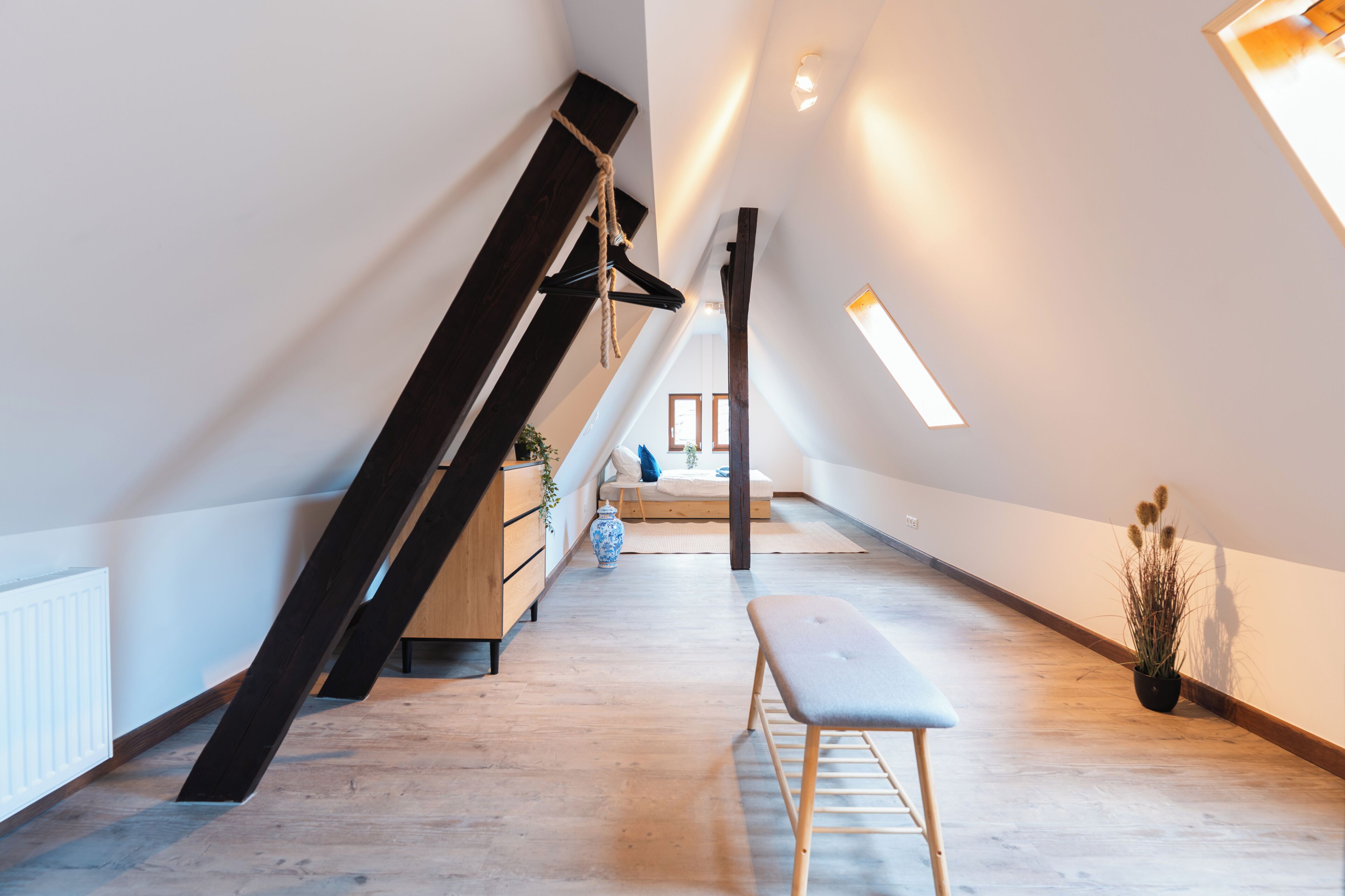 Een ruime zolderkamer met schuine wanden, een houten bankje, een klein tafeltje en een hangplant.