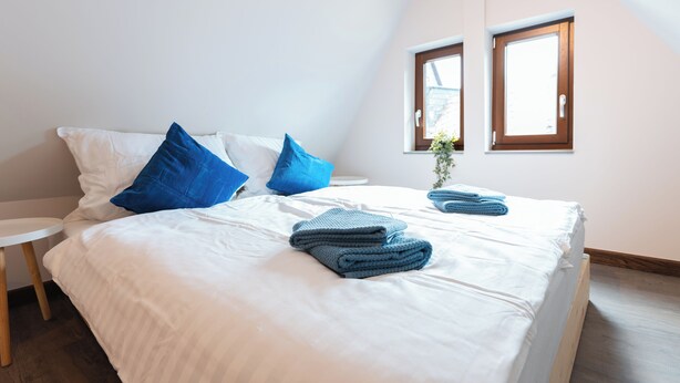 Una cama bien hecha con ropa de cama blanca y almohadas azules, dos mesitas de noche y dos ventanas con marcos de madera.