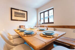 Een eetgedeelte met een houten tafel voor vier personen, een lijst met een foto aan de muur, en een raam met uitzicht op het omliggende landschap.
