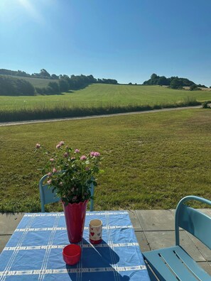 Ein Tisch mit blauer Tischdecke, ein roter Topf mit rosa Blumen, eine rote Tasse und eine Tasse mit Herzmuster an einem sonnigen Tag mit Blick auf ein grünes Feld und Hügel.