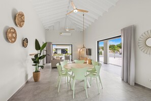 Ein moderner Essbereich mit einem runden Tisch und hellgrünen Stühlen, einem Deckenventilator und Blick ins Freie durch große Fenster.