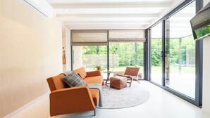 En moderne stue med sofa, skammel og lænestole, store vinduer og udsigt til grøn natur.
