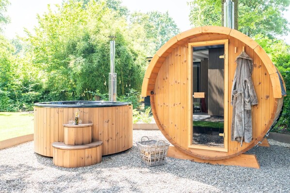 A wooden hot tub and a wooden cabin with a door open, set in a garden with trees.