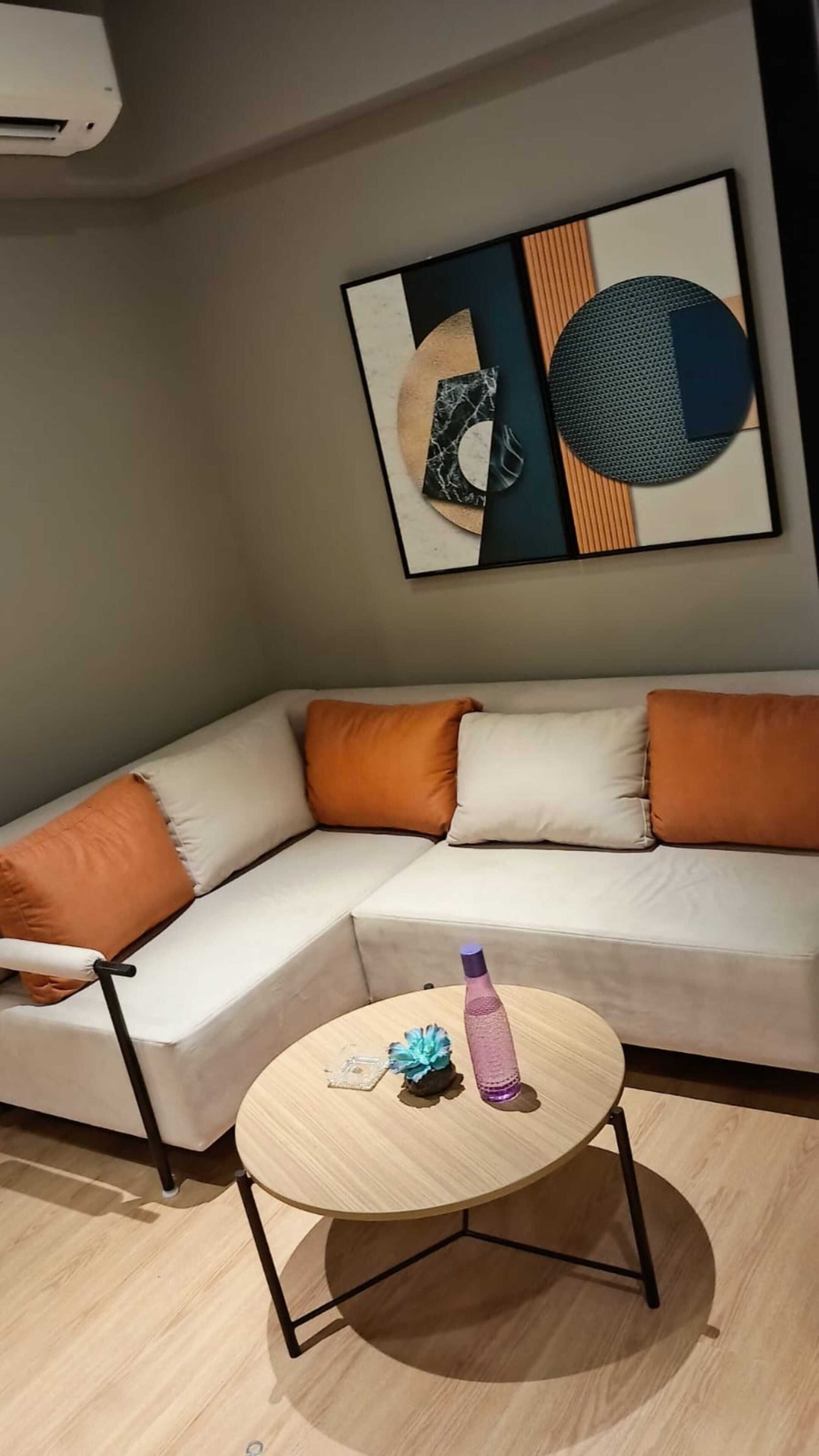 Een moderne woonkamer met een beige bank, oranje kussens, een ronde houten salontafel met een paarse waterfles, en een abstract kunstwerk in een lijst aan de muur.