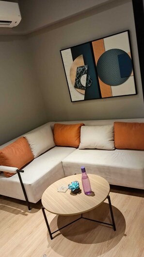 A modern living room with a beige sofa, orange cushions, a round wooden coffee table with a purple water bottle, and a framed abstract artwork on the wall. - Awesome The Studio Stay (Raipur)