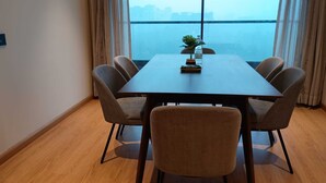 A dining area with a rectangular table, six chairs, a potted plant, and a view of a cityscape through large windows.