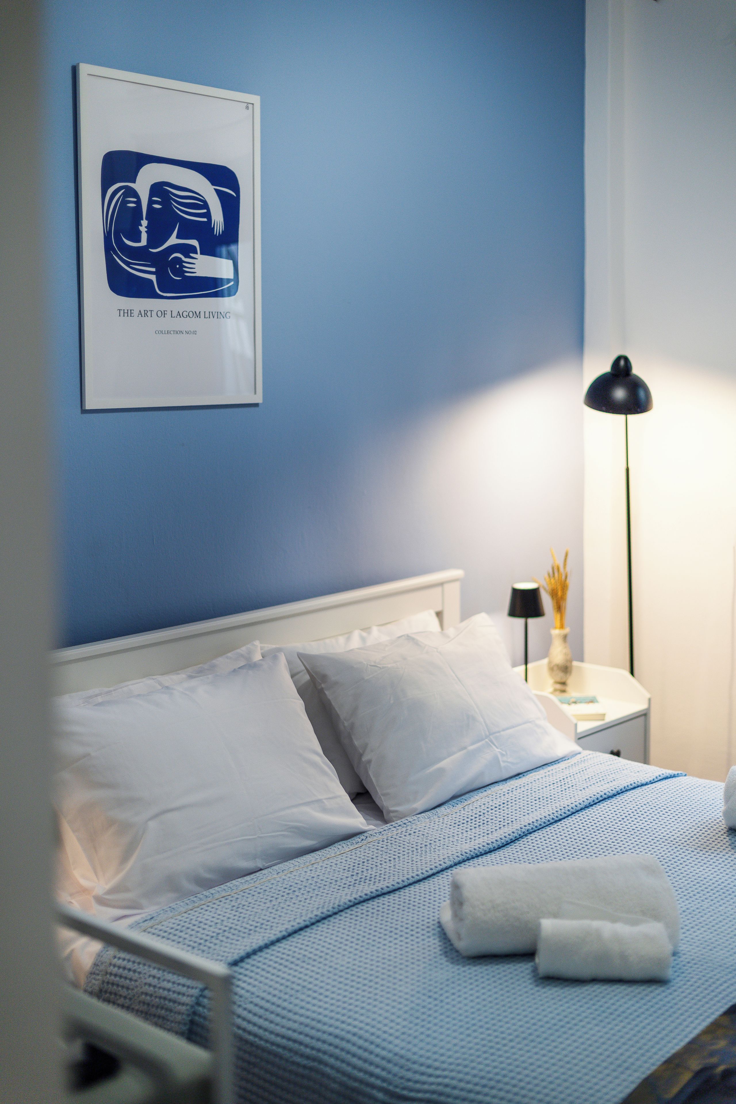 A neatly made bed with white linens and a blue patterned bedspread, a bedside lamp, and a framed artwork on the wall.