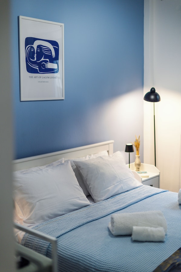 A neatly made bed with white linens and a blue patterned bedspread, a bedside lamp, and a framed artwork on the wall.