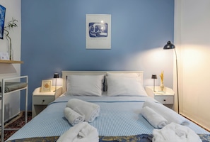 A hotel room with a bed, two bedside tables, a lamp, a framed artwork, and a small plant.