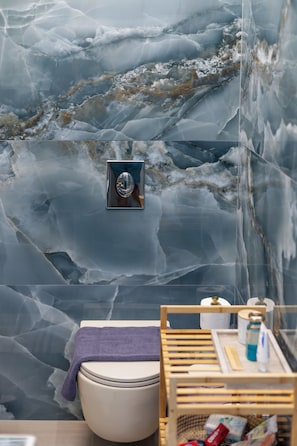 A bathroom with a marble wall, a toilet, a wooden shelf with toiletries, and a towel.