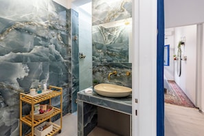 A bathroom with a marble wall, a wooden shelf with toiletries, a sink, and a mirror. - Διαμέρισμα στο κέντρο της πόλης. (Filippiada)