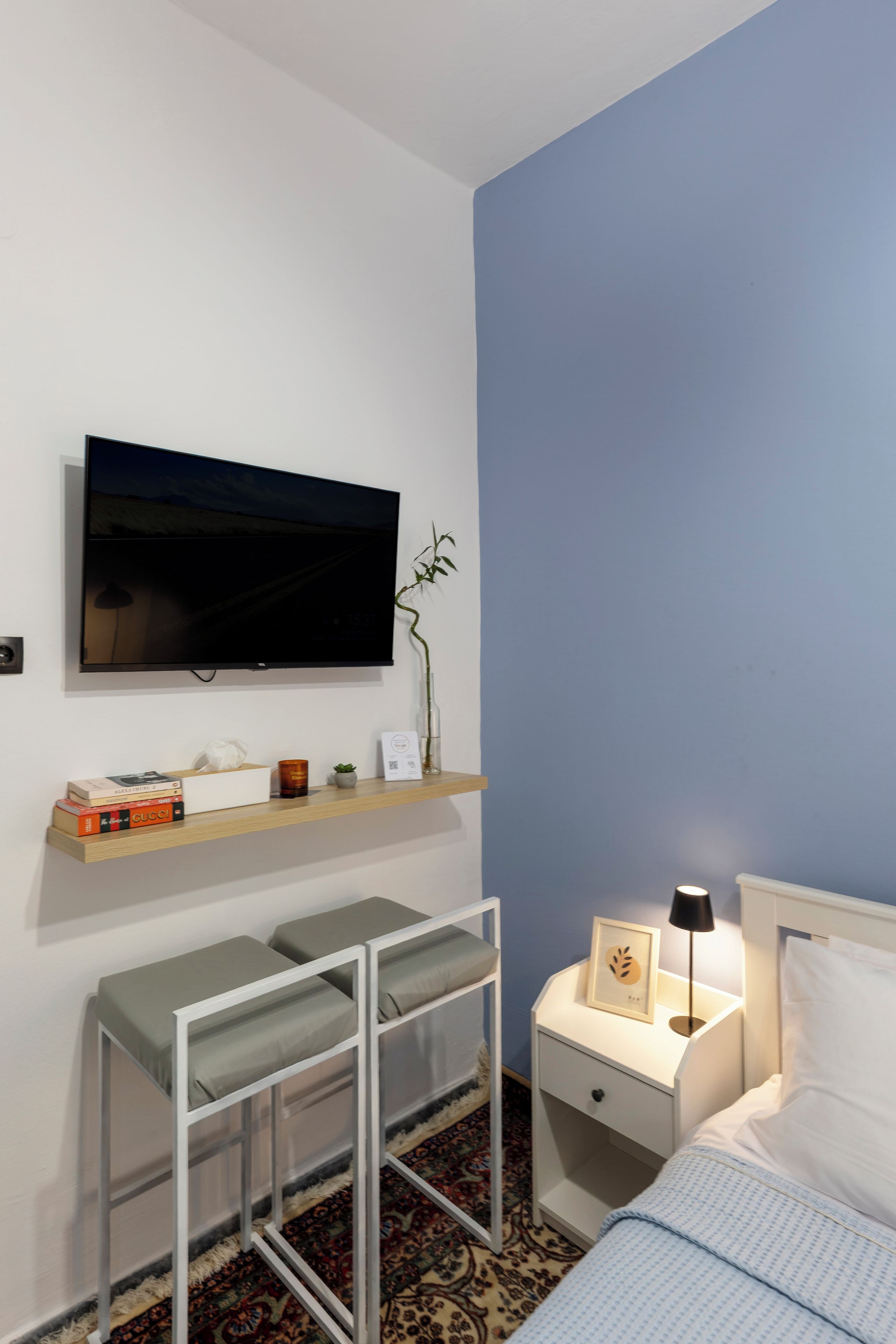 A bedroom with a bed, bedside table, TV, shelf with books, and a small plant.
