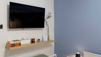 A bedroom with a bed, bedside table, TV, shelf with books, and a small plant.