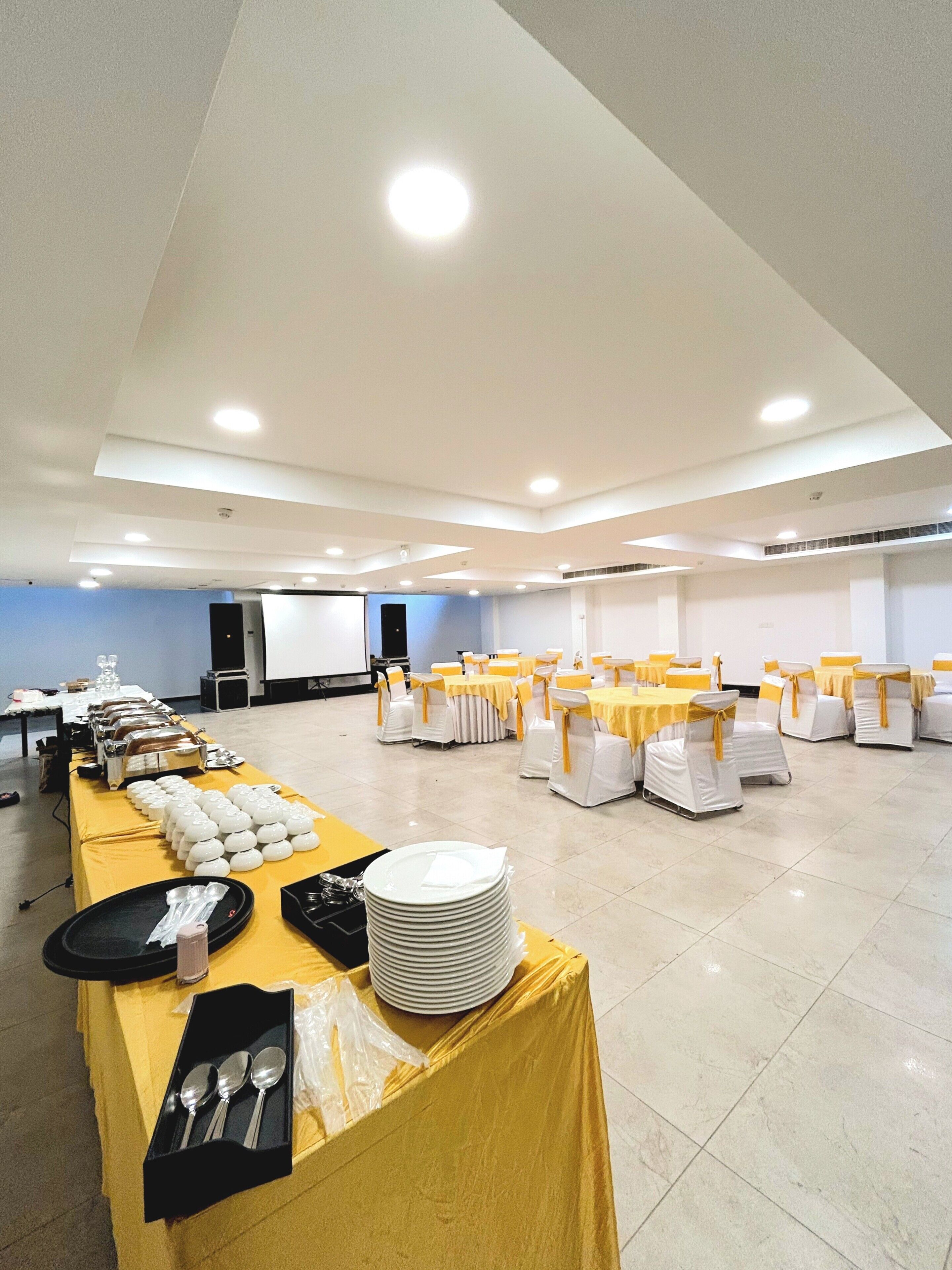 A spacious banquet hall with multiple round tables, each with white chairs and yellow sashes, and a buffet table with stacked plates and silverware.