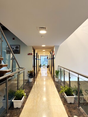 Un couloir d’hôtel moderne avec une rambarde en verre, des plantes en pot et un panneau indiquant la sortie de secours.
