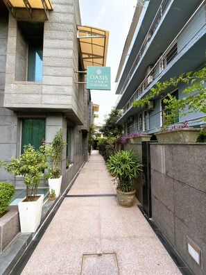 Un sendero estrecho flanqueado por edificios con macetas y un cartel que indica 'OASIS ONE GURUGRAM'.