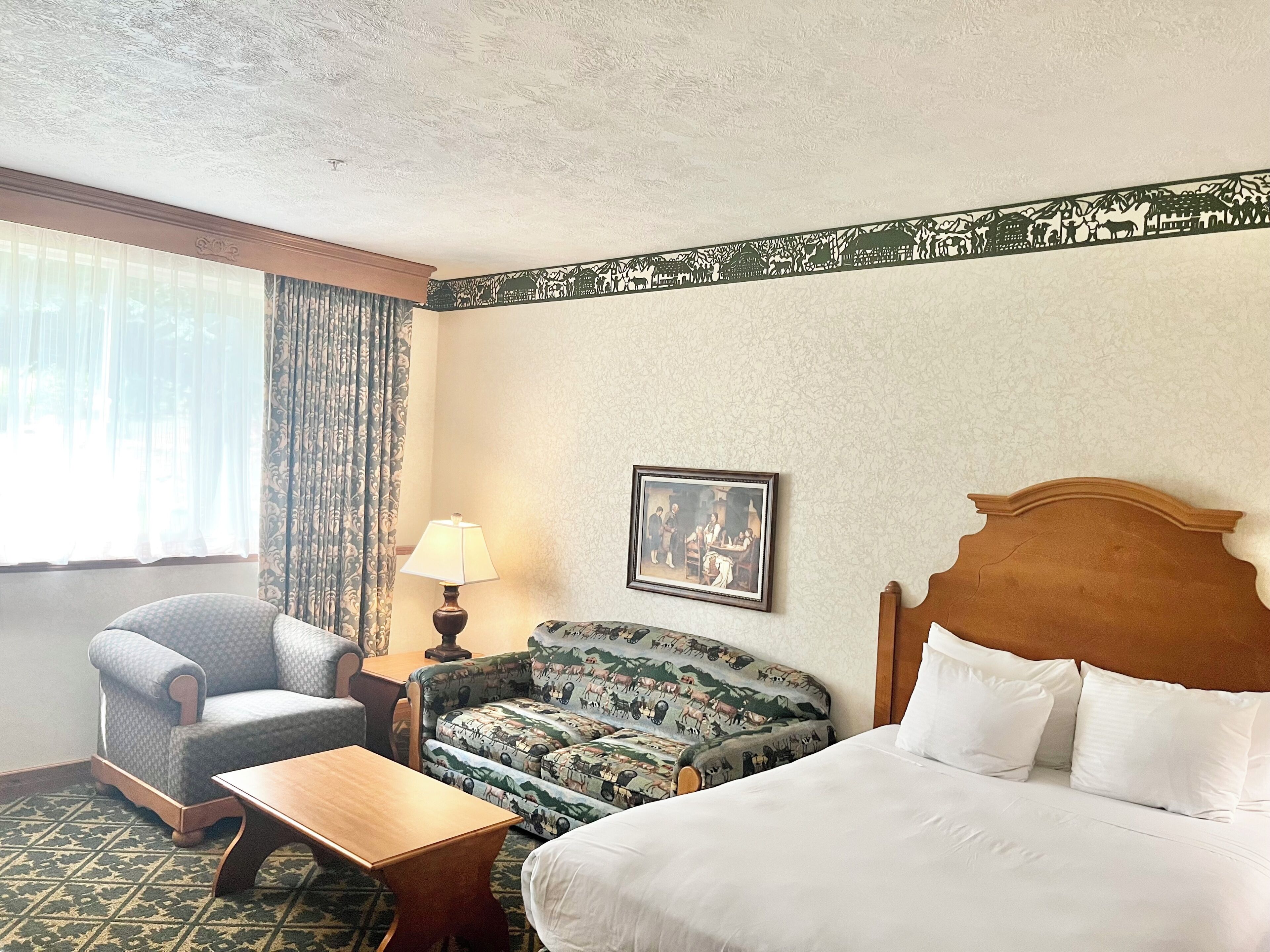 A hotel room with a bed, a chair, a sofa, a table, a lamp, and a painting on the wall.