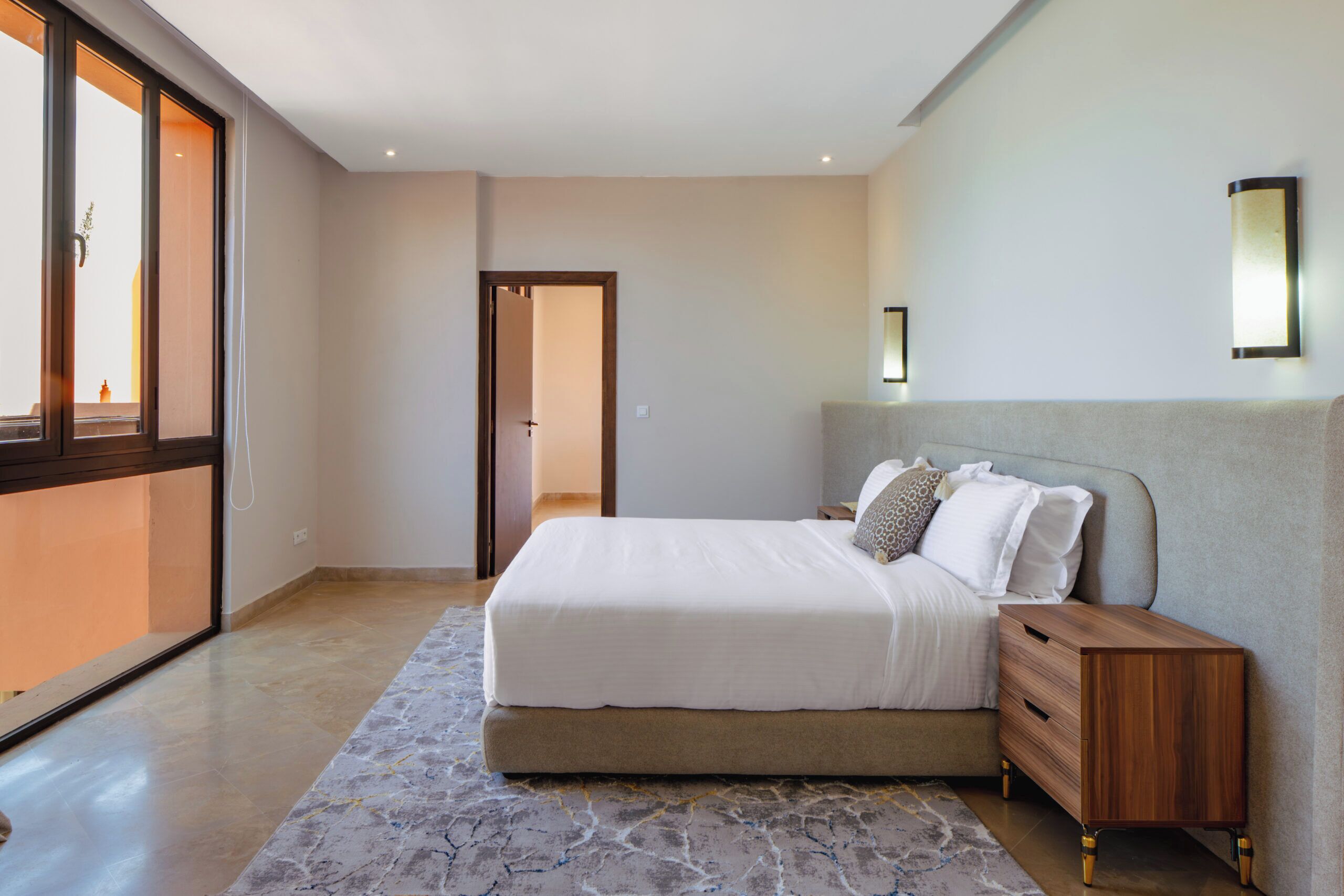 A modern bedroom with a large bed, a nightstand, a mirror, and a view through a large window.