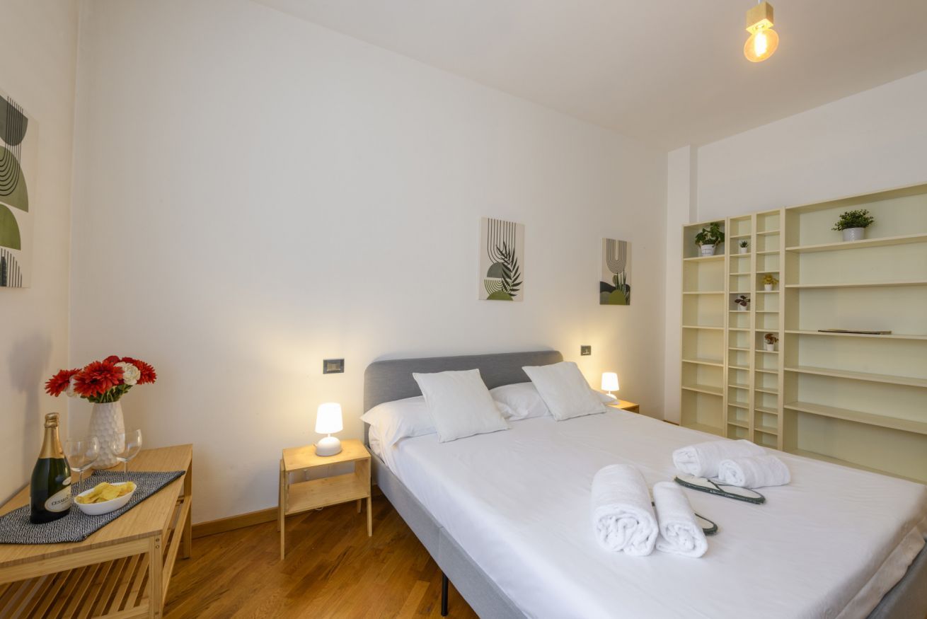 A hotel room with a bed, bedside tables, a nightstand, a small table with a bottle and glasses, and a shelf with plants.