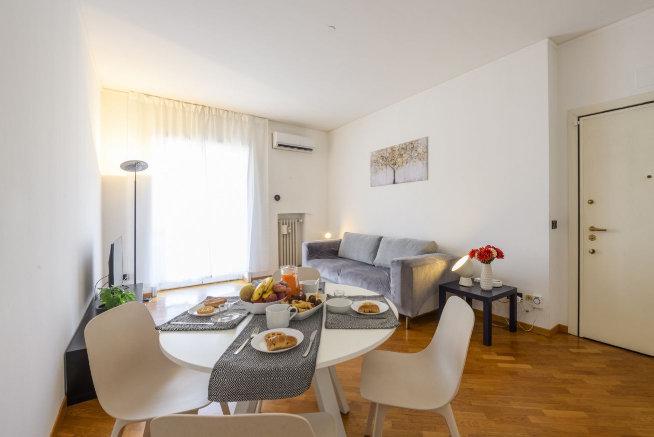 A modern living room with a dining area, a grey sofa, a small table with a lamp, and a door.