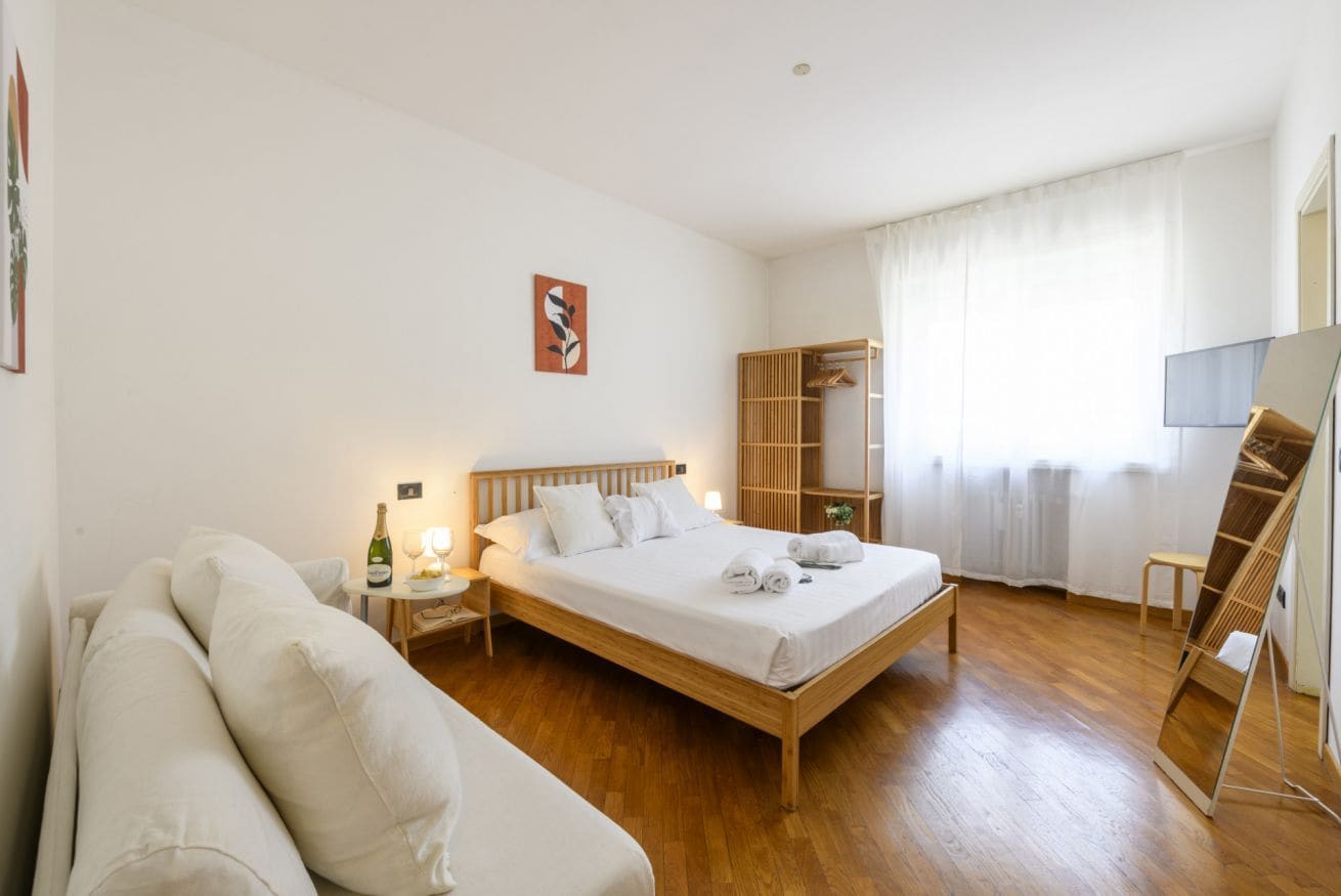 Een slaapkamer met een houten bed, een nachtkastje met een fles en glazen, een houten garderobe en een groot raam met gordijnen.