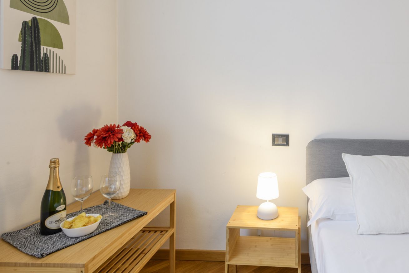 A hotel room with a bed, a bedside table with a lamp, a small table with a bottle and glasses, and a vase with flowers.
