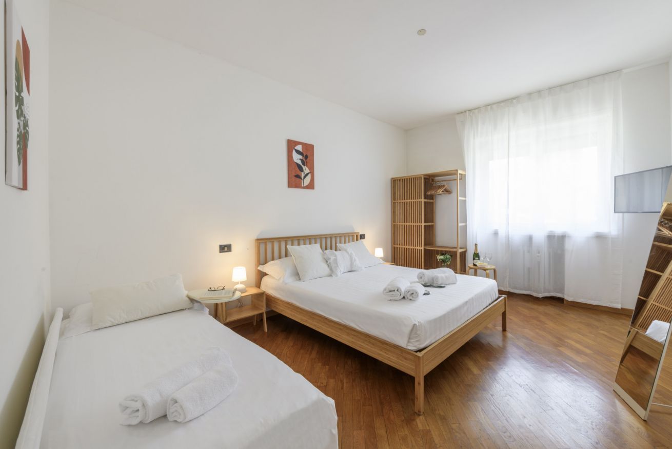 Habitación de hotel con dos camas, cabecera de madera, mesita de noche con lámpara, armario y una ventana grande con cortinas translúcidas.
