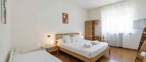 Habitación de hotel con dos camas, cabecera de madera, mesita de noche con lámpara, armario y una ventana grande con cortinas translúcidas.