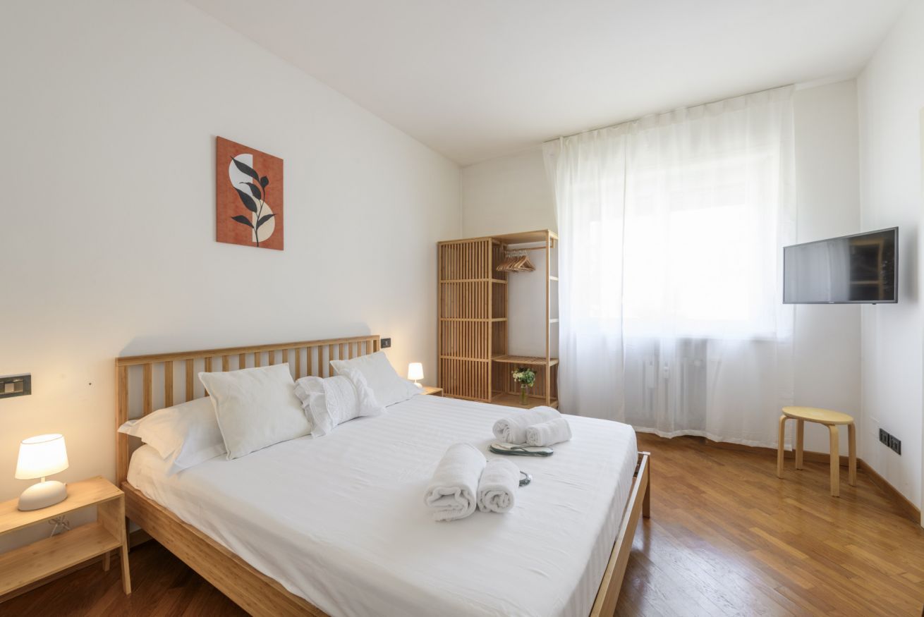 Een hotelkamer met een houten bed, een nachtkastje met een lamp, een aan de muur bevestigde televisie en een raam met dunne gordijnen.