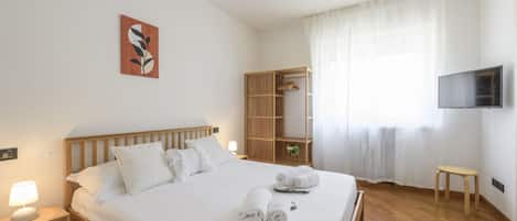 Habitación de hotel con una cama de madera, una mesita de noche con lámpara, un televisor montado en la pared y una ventana con cortinas traslúcidas.