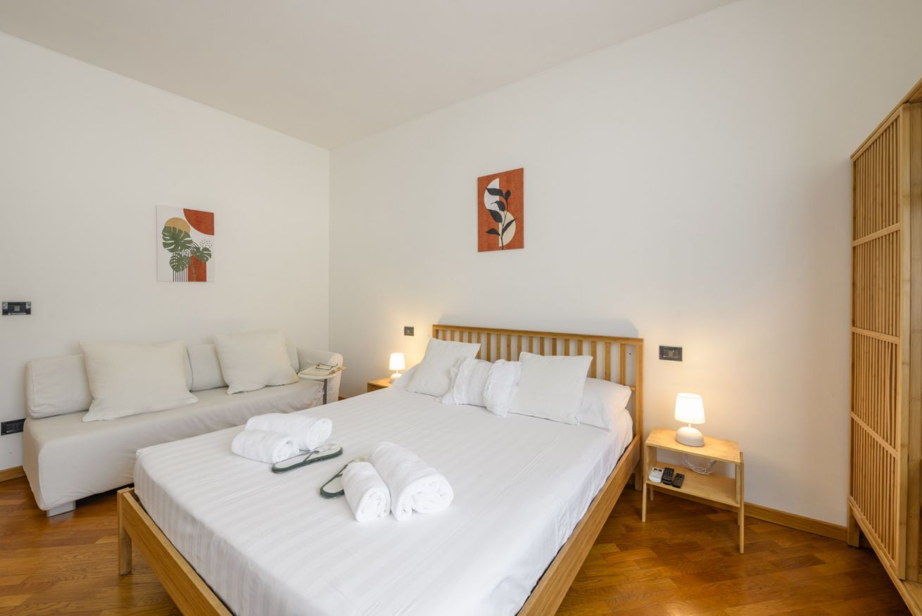 A hotel room with a double bed, a sofa, a bedside table with a lamp, and artwork on the walls.