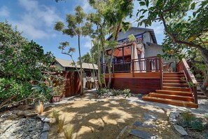 A residential property with a wooden staircase leading to a balcony, surrounded by lush greenery and a stone pathway. - Moden Craftsman • Pet Friendly • Sleeps 6 (Long Beach)