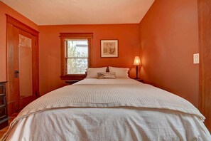 Une chambre à coucher avec un grand lit, une fenêtre avec des stores, une lampe de chevet et une porte.