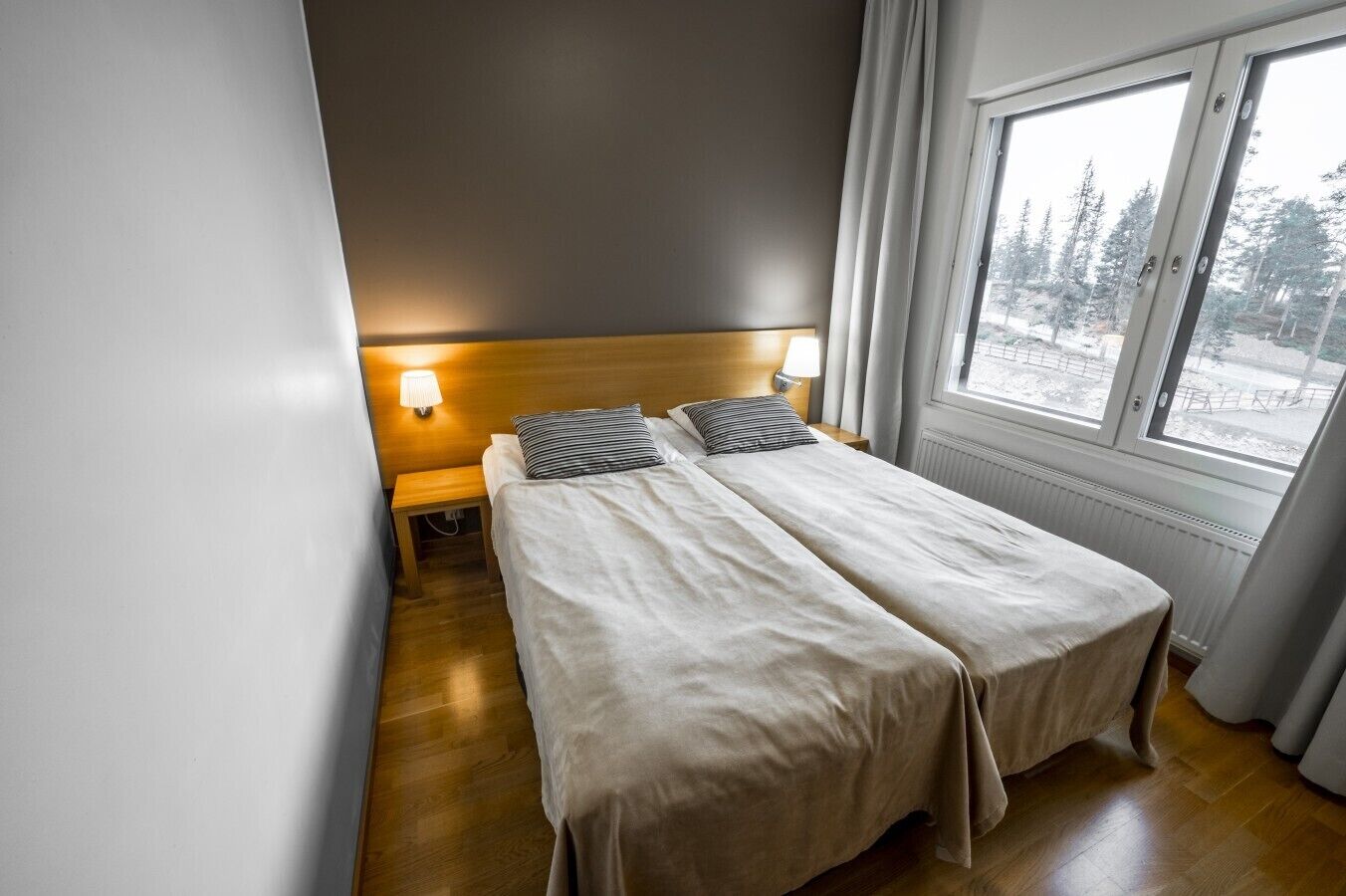 A hotel room with two beds, a wooden headboard, bedside lamps, and a window with a view of trees.