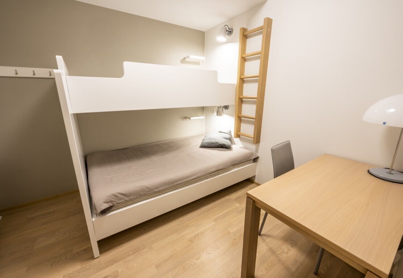A bunk bed with a ladder, a desk, and a lamp in a room with wooden flooring.