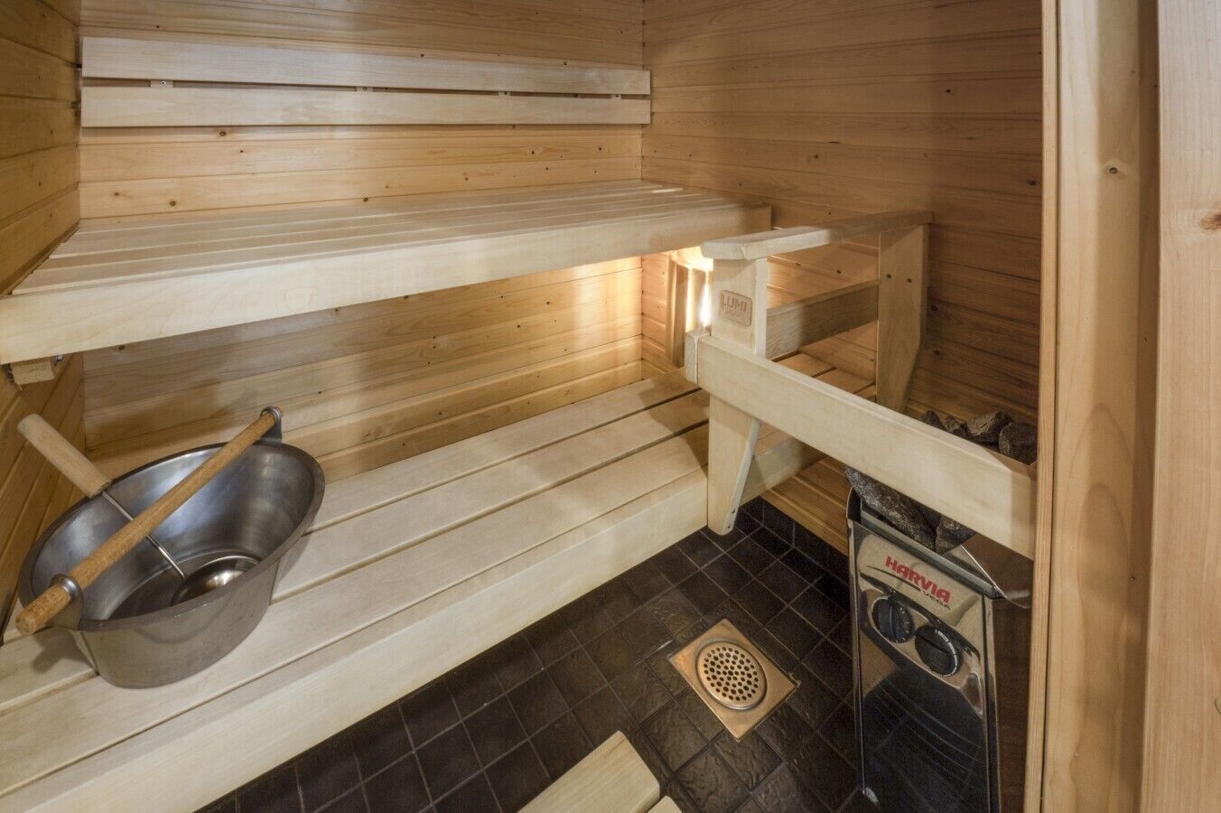 A sauna room with wooden benches, a metal basin, and a sauna heater.
