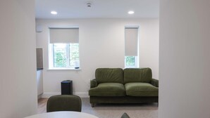 A room with a green sofa, a white table, and two windows with blinds. - The Maycliffe - Studio near Torquay Harbour (Torquay)