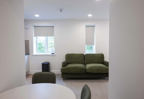 Ein Zimmer mit einem grünen Sofa, einem weißen Tisch und zwei Fenstern mit Jalousien.