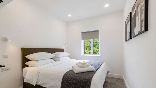 Cama bem arrumada com lençóis brancos e cabeceira escura em quarto com paredes brancas e janela com vista para o verde.