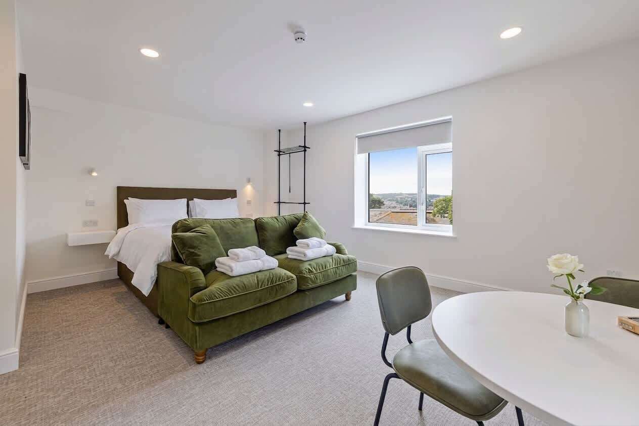 Une chambre avec un lit, un canapé vert, une table à manger ronde et une fenêtre donnant sur un beau paysage.
