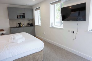 A modern hotel room with a bed, a TV mounted on the wall, a kitchen area, and a window providing natural light. - The Maycliffe - Modern Studio nr Harbour (Torquay)