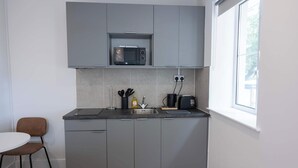 Eine kompakte Küche mit grauen Schränken, einer Mikrowelle, einem Wasserkocher und einem Toaster. Links befindet sich ein kleiner Tisch und ein Stuhl.
