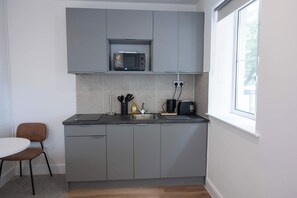 A compact kitchen with grey cabinets, a microwave, a kettle, and a toaster. There's a small table and a chair to the left. - The Maycliffe - Modern Studio nr Harbour (Torquay)