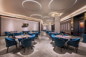 A modern dining area with round tables and blue chairs, featuring a central light fixture and a wall-mounted mirror. - Orange Crystal Hotel, Beijing Capital International and Exhibition Center (Shunyi)
