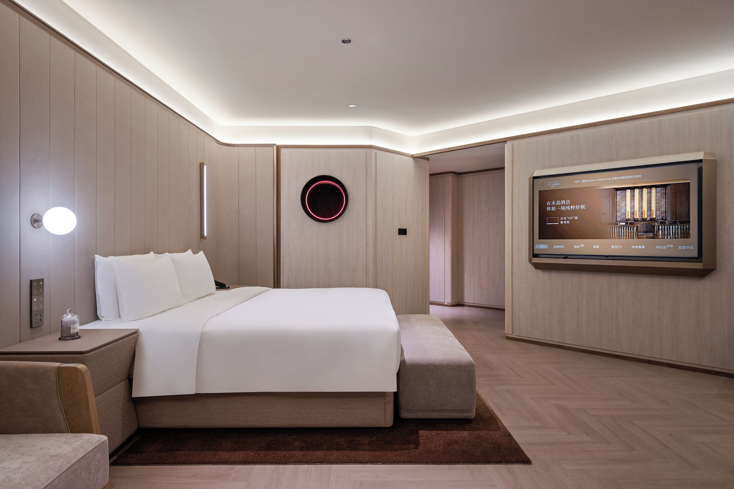 A modern hotel room with a large bed, bedside tables, a flat-screen TV, and a wall-mounted circular light fixture.