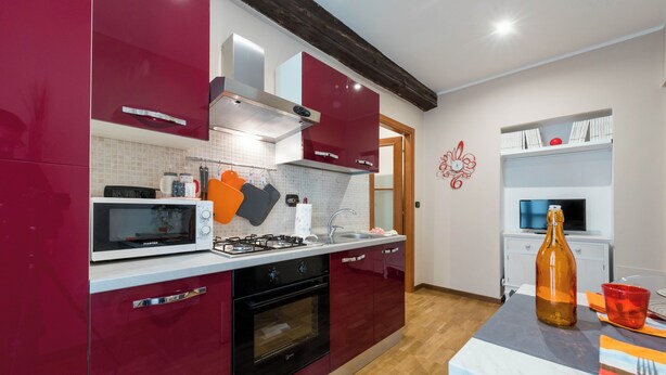 Een moderne keuken met rode kastjes, een magnetron, fornuis en een spoelbak. Er staat een tafel met een fles en een beker.