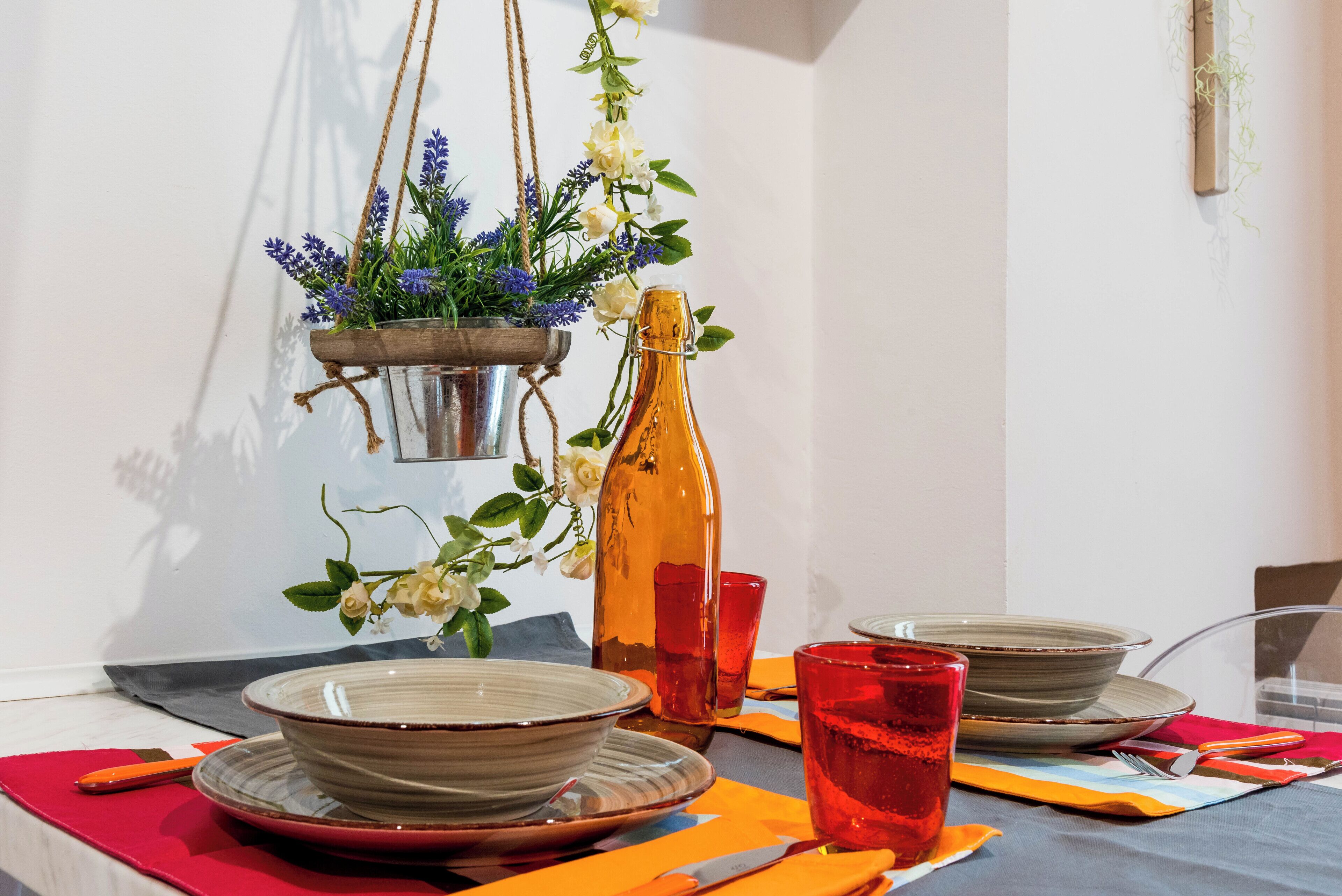A dining table with plates, bowls, and a bottle, set with orange napkins and red glasses.