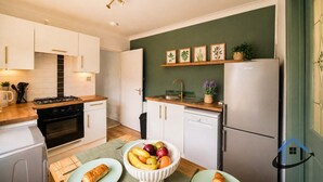 A compact kitchen with a gas stove, oven, and dishwasher. There is a dining table with plates and a bowl of fruit. - Barret Flat, sleeps 4, central location, Penarth (Penarth)
